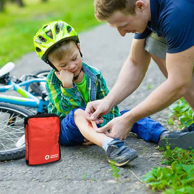 A parent applying a bandage from a Red Cross first aid kit to a child's shin.
