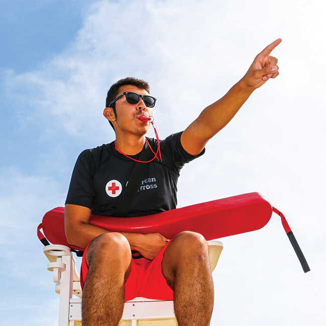 A lifeguard on duty signaling with a whistle while sitting in a lifeguard chair and holding a rescue tube.