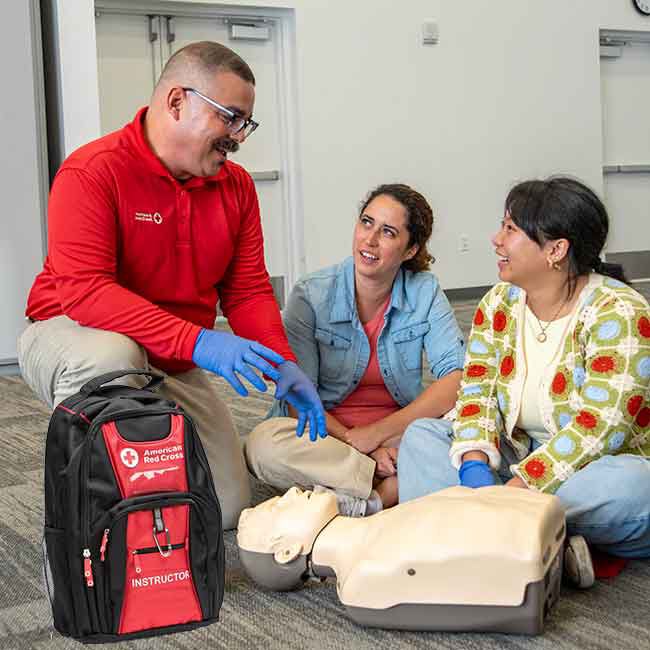 Red Cross CPR instructor talking to two students next to a CPR manikin and Red Cross Instructor Backpack.