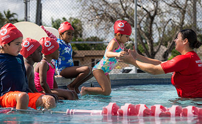A group of kids sitting at the edge of a pool and one student jumping into the water with a Red Cross Swimming Instructor.